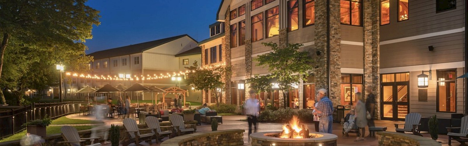 Outdoor gathering area with a fire pit and seating, part of a large building. String lights illuminate the space at dusk.