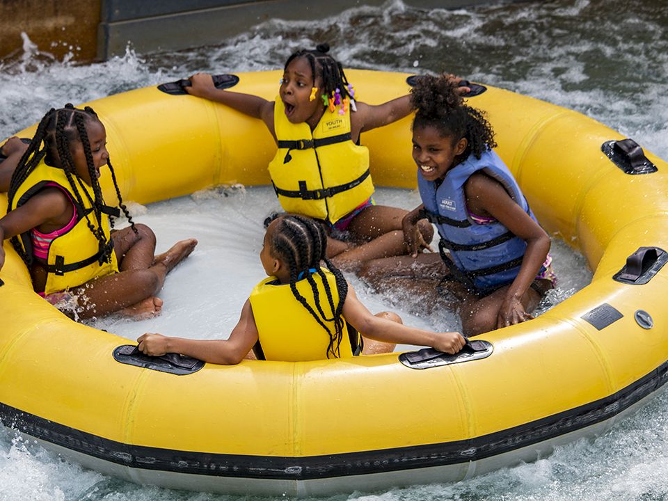 A group of kids in bright life jackets rides together on a yellow inflatable raft in choppy water, smiling and splashing as they float.