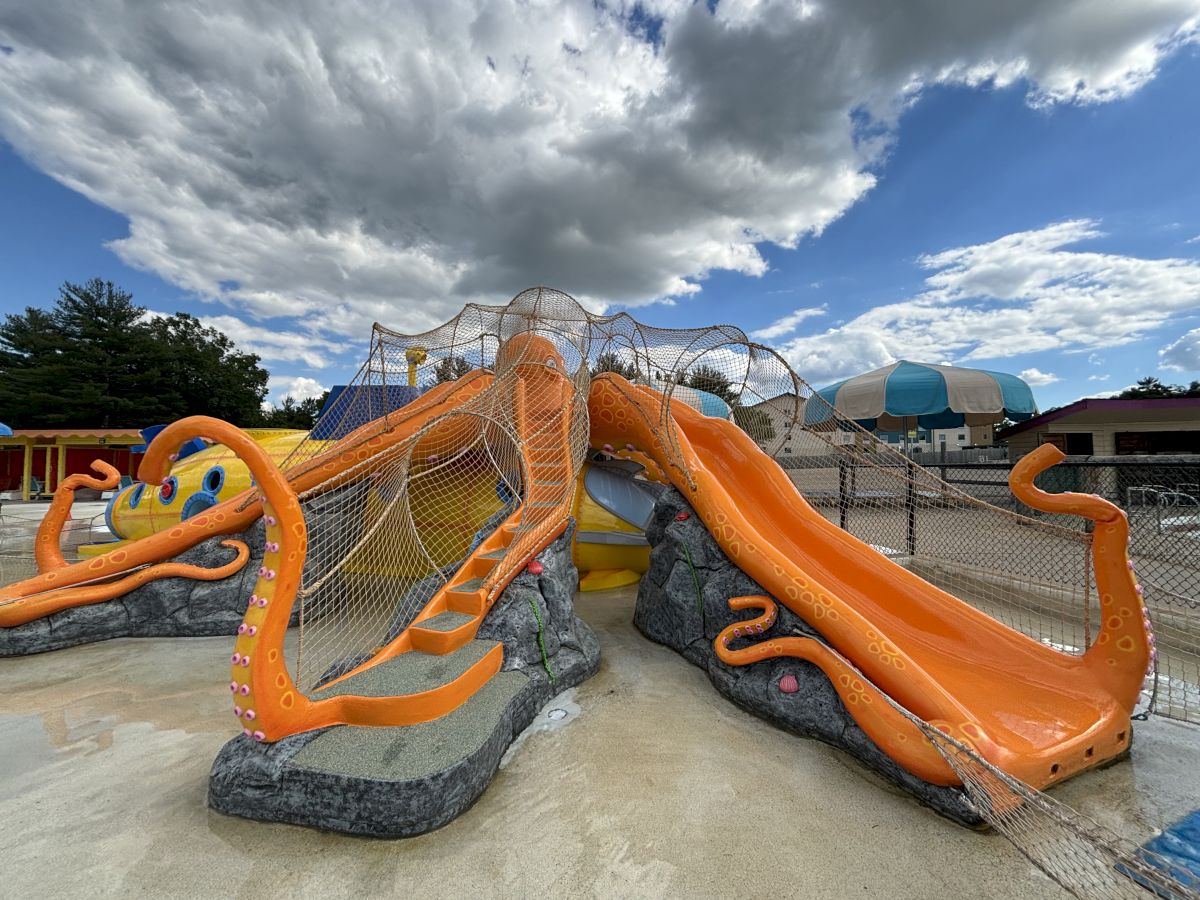 An outdoor playground with orange animal-themed slides inside a netted play area under a cloudy, blue sky.