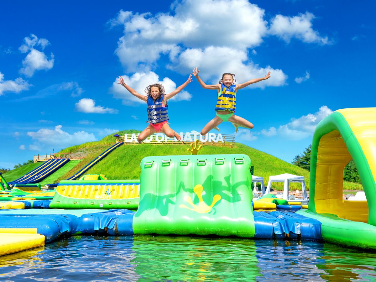 Two kids in life jackets joyfully jump from a bright inflatable water slide, splashing in a backyard splash park under sunny clouds.