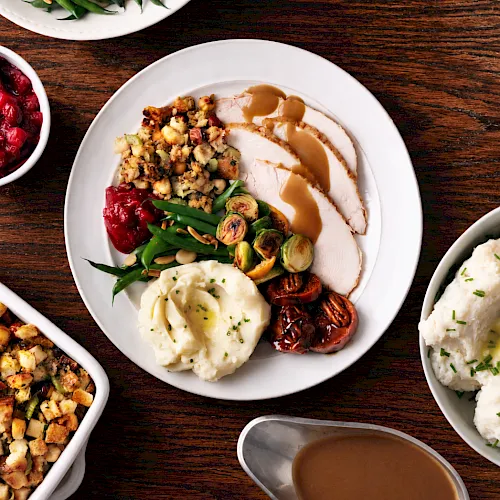 A plate of sliced roast turkey with brown gravy, mashed potatoes, and roasted brussels sprouts, plus green beans and cherries.