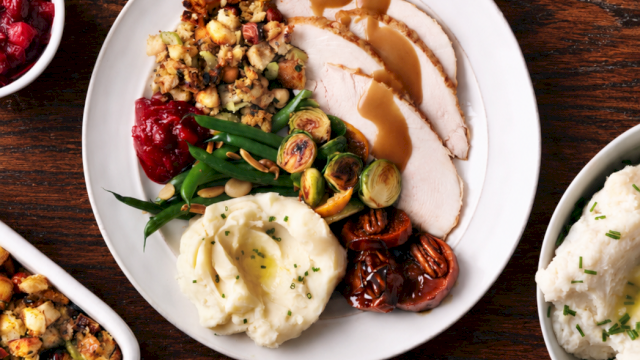A plate of sliced roast turkey with brown gravy, mashed potatoes, and roasted brussels sprouts, plus green beans and cherries.