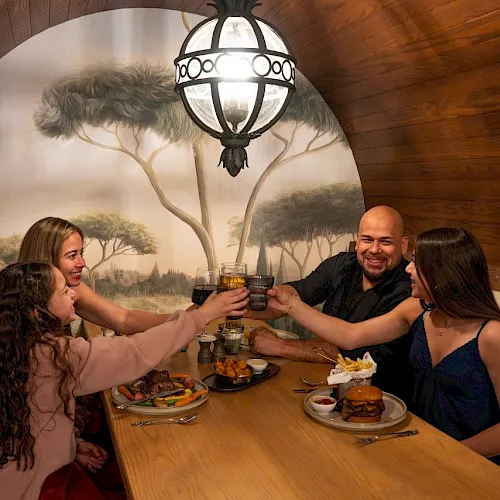 A group of friends cheers with drinks at a restaurant table, smiling under warm lights and a nature-themed mural.