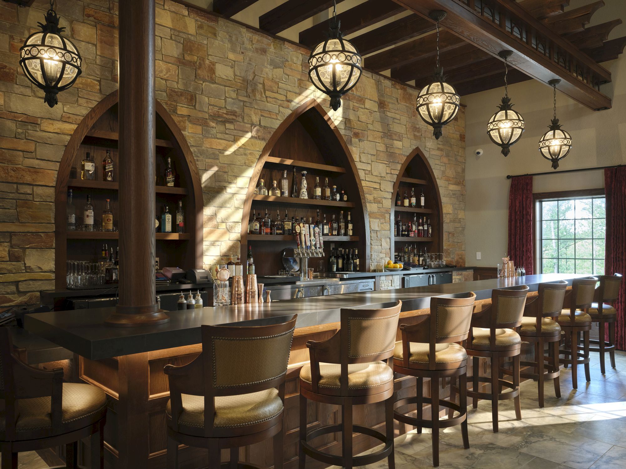 A cozy bar with stone arches, dark wood shelves, row of stools, and warm hanging lanterns; bottles line the back wall near a sunny window.