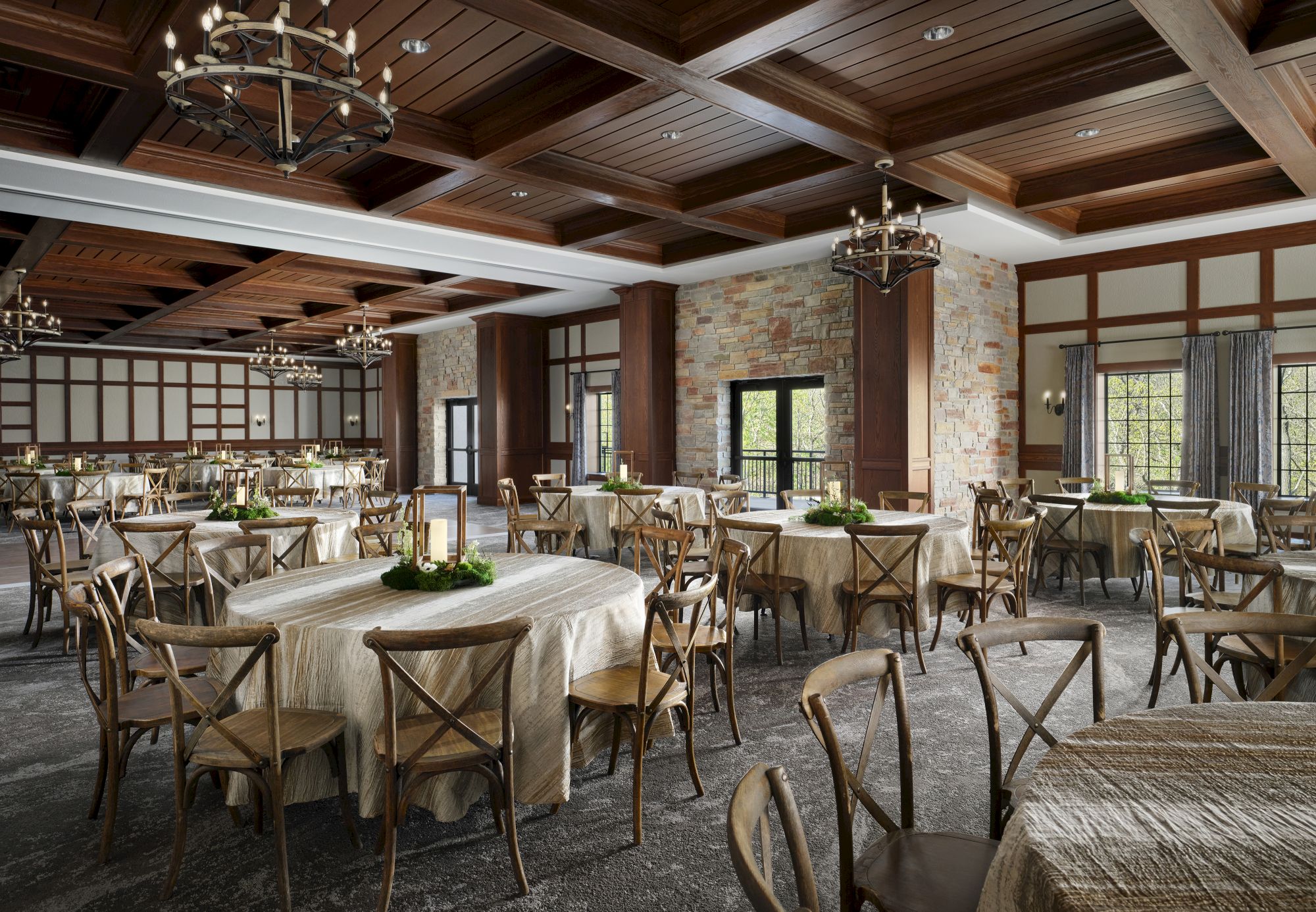 Rustic banquet hall with wood beams, brick walls, and multiple round tables set for an event; warm lighting and elegant chandeliers abound.