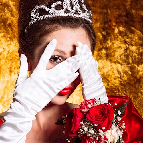 A person in a red dress and white gloves, wearing a tiara, hiding part of their face with hands, holding a bouquet of red roses.