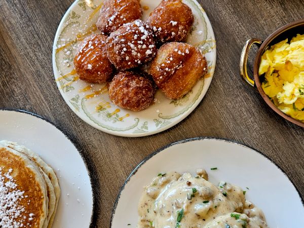 A table full of comfort food: croissants, fried dough bites with glaze, buttered biscuits, creamy chicken with herbs, and a side dish with scrambled eggs or potatoes, all arranged on plates.