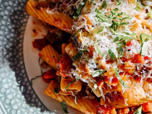 A plate of loaded penne pasta with tomato sauce, grated cheese, and fresh herbs piled high on a decorative bowl. (Note: ends with a period.)