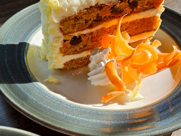 A slice of carrot cake with cream cheese frosting, layered with orange ribbons, on a plate with whipped cream d&eacute;cor, beside a dessert plate.