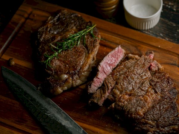 Juicy grilled steak slices on a wooden board with a herb garnish and a bread knife nearby.