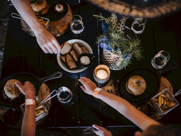 A top-down dinner scene with hands reaching for food, bowls of bread and fries, candles, and a rustic table setup for a shared meal.