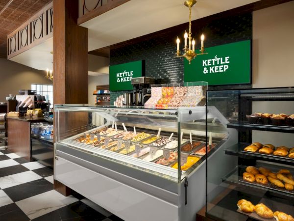 A bakery display with assorted pastries and donuts in a glass case, shelves of bread, and a checkered floor in a cozy cafe setting.