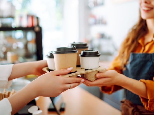 Two hands pass a set of takeout coffee cups to a barista in a cozy coffee shop, with a warm, friendly vibe.