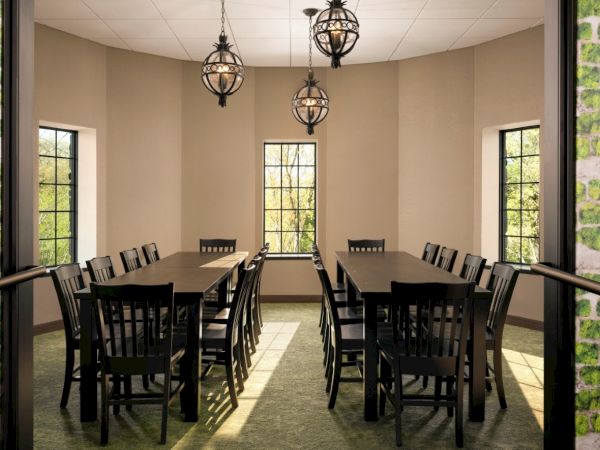 A small dining room with two long wooden tables and matching chairs, pendant lights, beige walls, and windows letting in daylight.