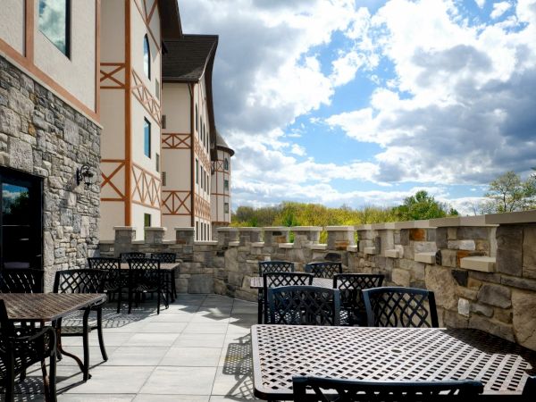 Outdoor patio with metal lattice tables and chairs, stone wall and building, blue sky with clouds, daylight, resort-like ambiance. End.
