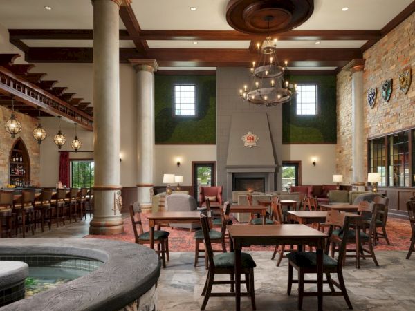 A spacious, upscale restaurant with wooden tables and chairs, a central fireplace, stone walls, chandeliers, and a warm, inviting interior.