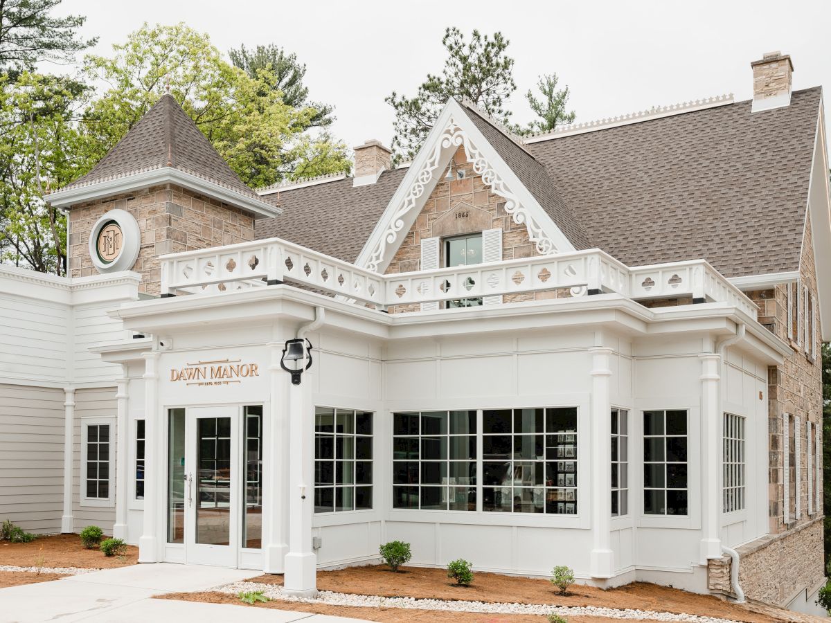 A charming white storefront with a gabled roof, large windows, and classic details, nestled among trees and a light entrance path.