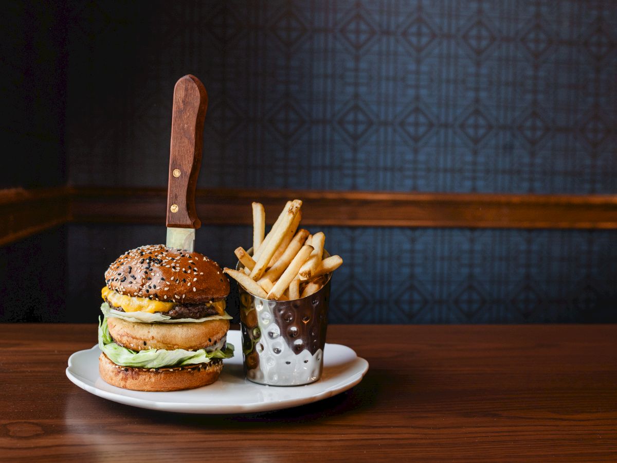 A burger with lettuce and cheese on a plate, a side of French fries in a metal cup, and a large knife stuck into the bun, on a wooden table.