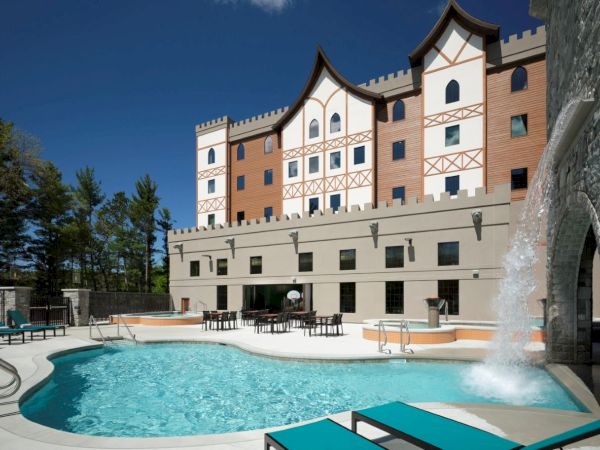 A resort with a curved outdoor pool, turquoise water, sun loungers, and a beige-and-brown castle-like building with arched windows and a waterfall fountain.