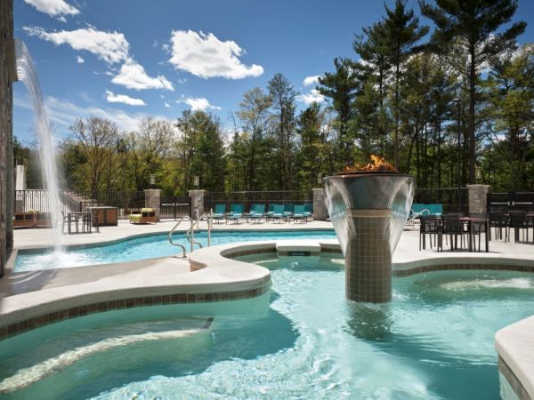 A luxurious pool area with a hot tub, water features, lounge chairs, and lush trees under a bright, partly cloudy sky, perfect for relaxation.
