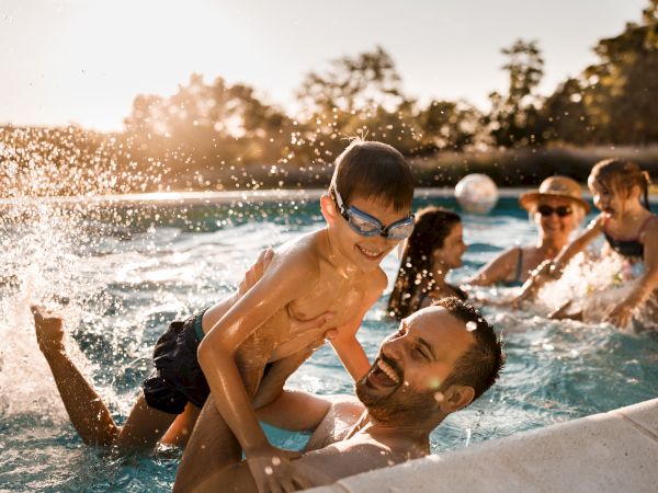 A man and child playfully splash and ride in a sunny pool with friends cheering in the background, smiling and enjoying a summer day.