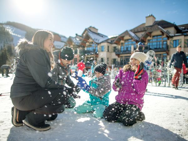 A group of kids and an adult play in the snowy town square, throwing snow and laughing in bright winter light.