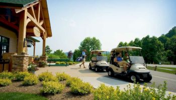 People near a building, with two golf carts parked on the path. One person holds a golf bag as greenery surrounds the area.