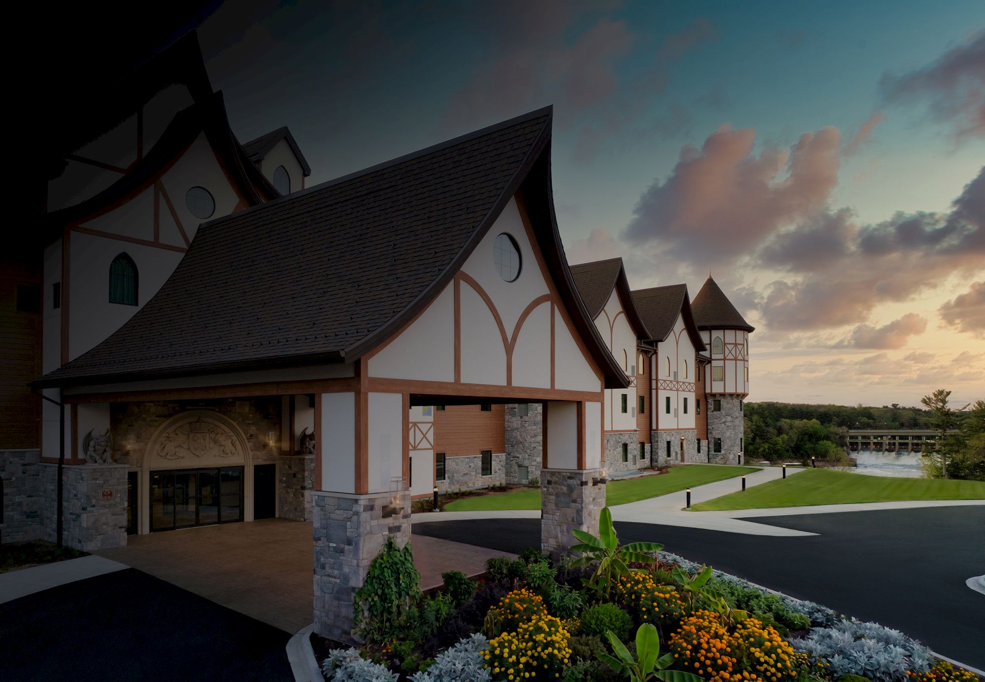A picturesque half-timbered mansion with arches and manicured gardens along a walkway, set under a colorful sunset sky.