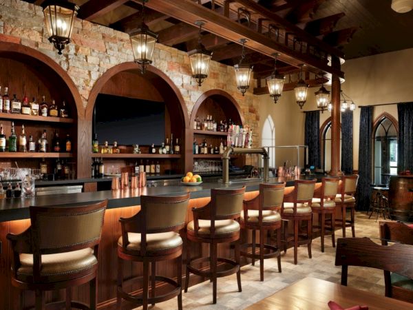 A rustic bar interior with wood arches, hanging lanterns, and a row of leather stools at a curved counter lined with bottles and glasses.