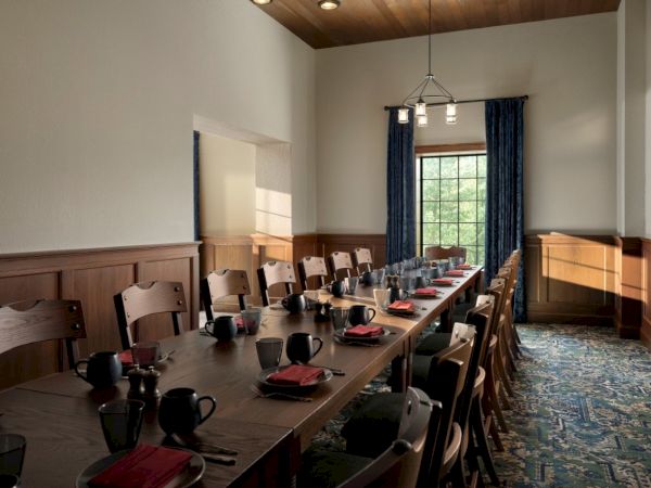 A long dining table set with plates, glasses, and utensils in a cheerful room, wooden chairs along both sides, natural light from a window.