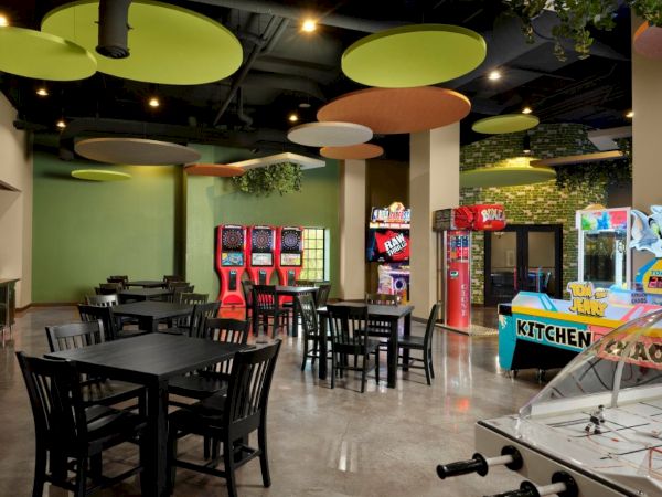 A bright game room or cafe with black tables and chairs, colorful circular ceiling panels, arcade machines, and a foosball table.