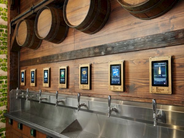 Tap handles, wooden barrels, and digital menus on a rustic wall&mdash;self-pour taps line a stainless sink, with glowing screens above each tap.
