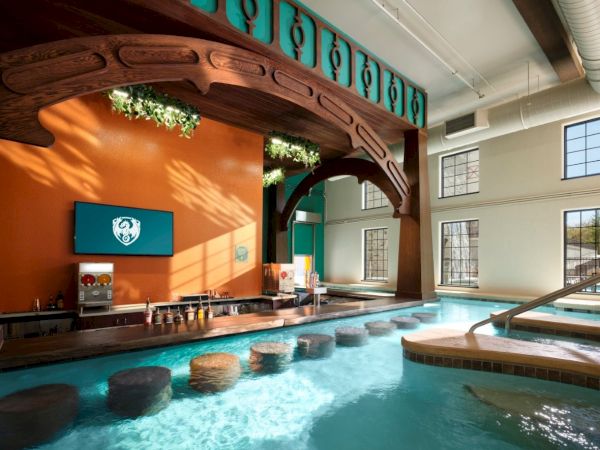 Indoor resort pool area with orange wall, wooden archway, teal accents, sunlit windows, and row of stepping stones across the pool.