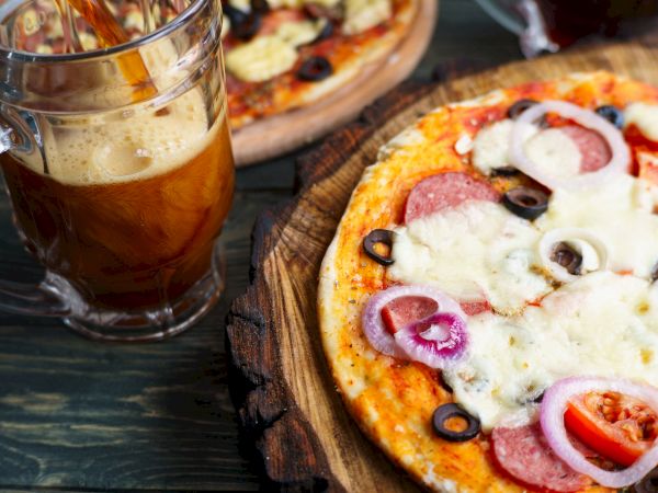 A pizza with cheese, pepperoni, olives, onions, tomatoes on a wooden board alongside a glass mug of coffee; casual outdoor dining.
