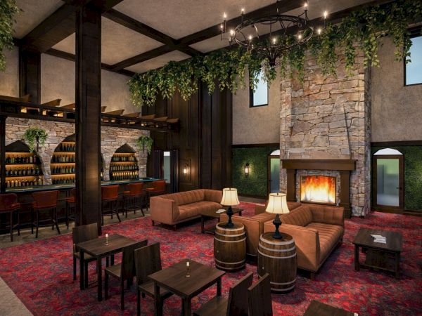Cozy rustic lounge with a stone fireplace, leather sofas, dark wood beams, hanging greenery, and a bar area with barrels and warm lighting.