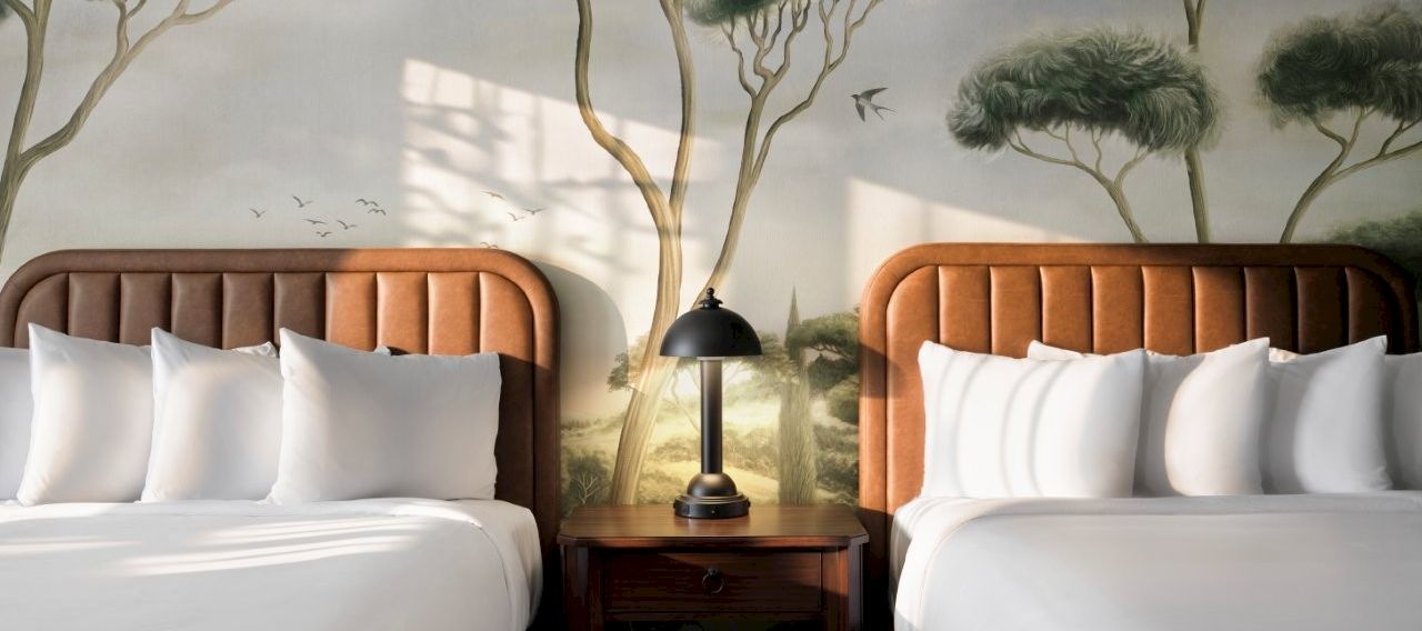 A cozy bedroom scene with two made-up beds, a wooden nightstand, a black lamp, and a nature forest mural on the wall.