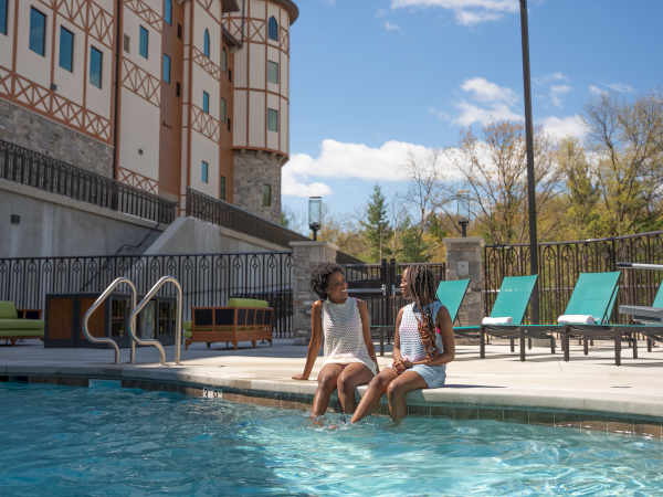 Two women chat and smile by a sunny outdoor pool at a resort, with cabanas and loungers in the background.