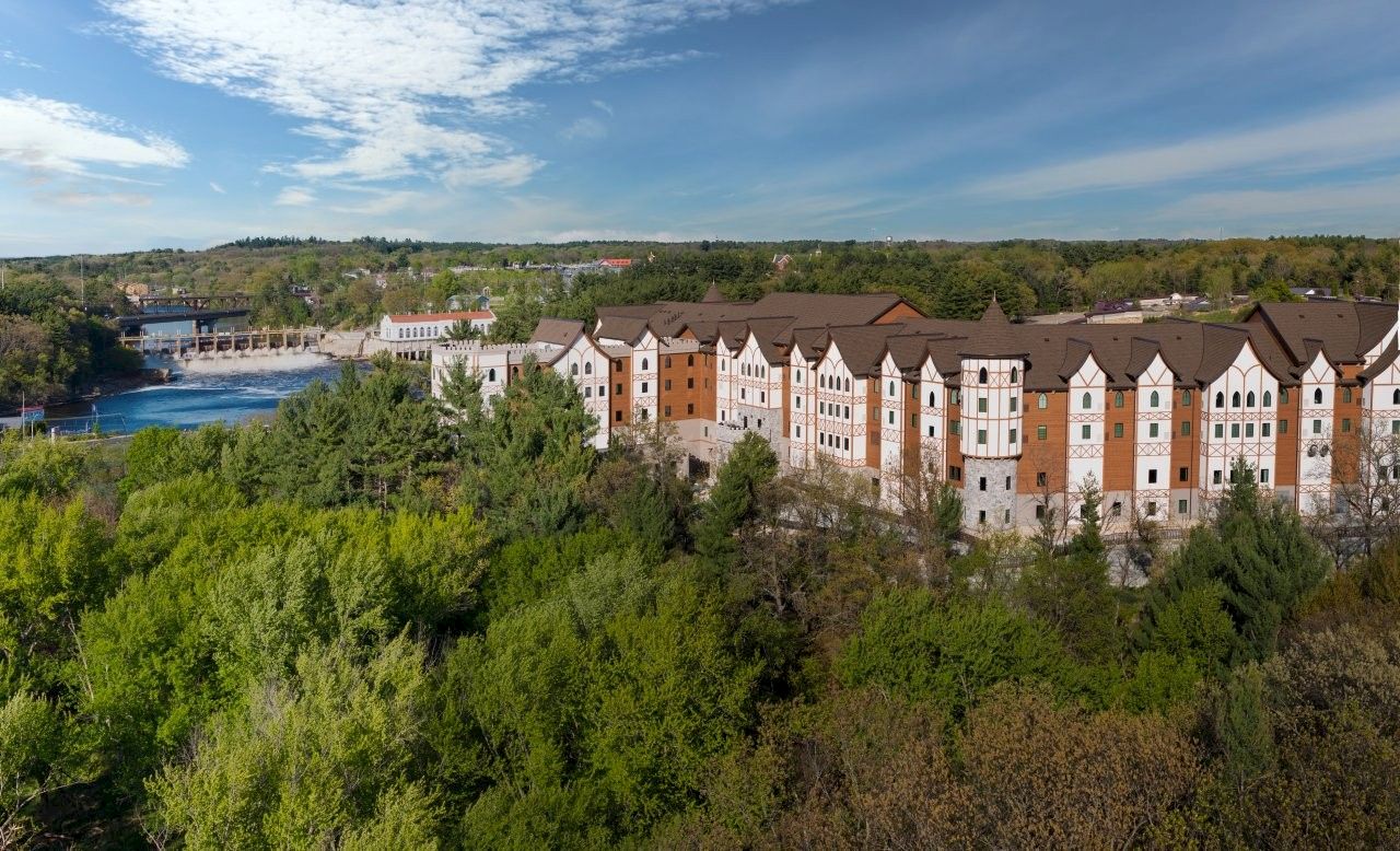A riverside hotel complex with colorful half-timbered buildings and turrets, surrounded by lush trees under a blue sky.