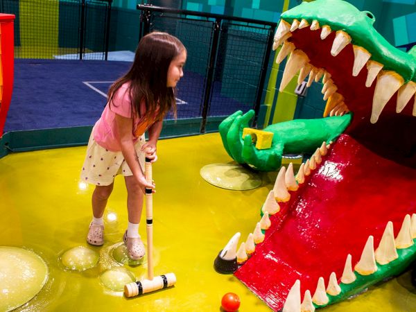 A girl in a pink shirt and white skirt plays mini golf, aiming at a large green dinosaur mouth with sharp teeth.