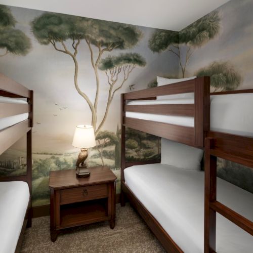 A small dorm-style room with two wooden bunk beds, a nightstand, and a lamp, all against a mural of trees and a scenic wall.