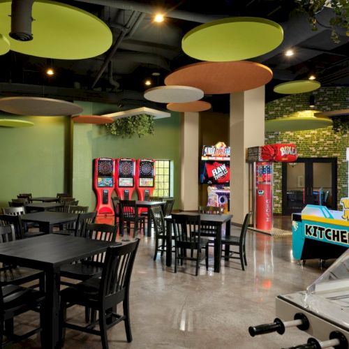 A bright arcade-style cafe area with black tables and chairs, colorful ceiling disks, a green accent wall, vending machines, and a foosball table in a game zone.
