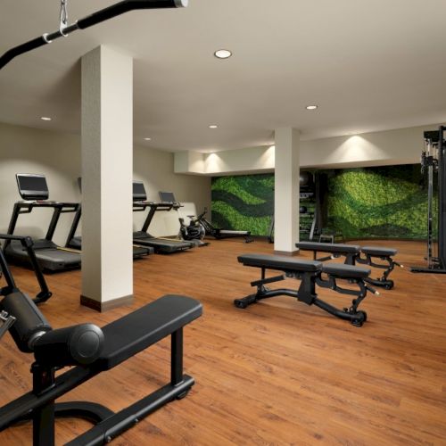 A modern gym with wooden floors, treadmills, benches, and a cable machine, bright with recessed lights and green wall murals.
