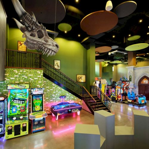 A bright arcade/play area with neon arcade games, a green-tilled wall, a staircase, seating blocks, and colorful ceiling decor.