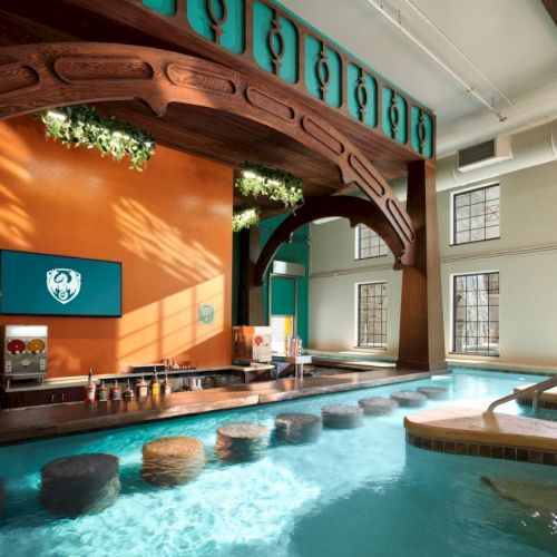 Indoor pool area with a bright orange accent wall, wooden arch decor, swim-up seating, large windows, and a hot tub.