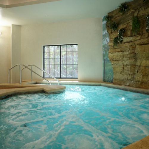 Indoor spa pool with a rock wall, bubbly turquoise water, and metal handrails by the steps, cozy lighting, and windows open to nature.