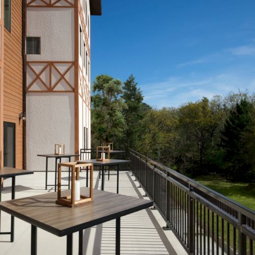 An outdoor balcony with wooden tables and lanterns, overlooking green trees under a clear blue sky, attached to a brick-and-wood building.