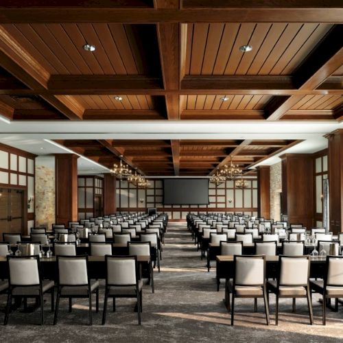 A large conference room with rows of chairs facing a central stage and screen, wood-paneled ceiling, chandeliers, and stone walls, ready for a presentation.