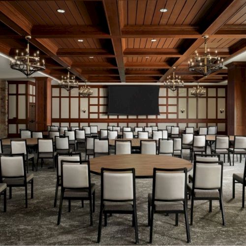 A conference room setup with rows of chairs facing a stage and big screen, brick walls, wooden ceiling panels, and chandeliers.