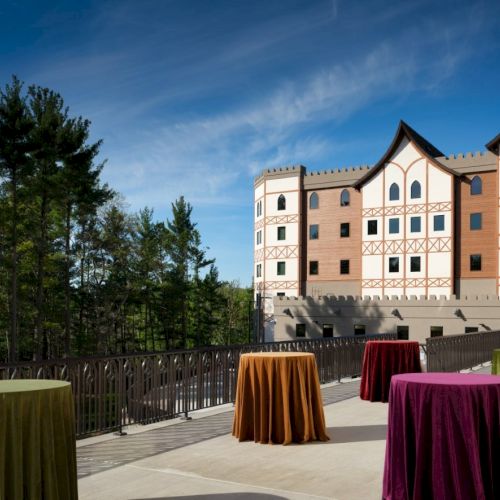 Colorful round tables covered in velvet on a terrace with a grand castle-like building in the background, under a blue sky.
