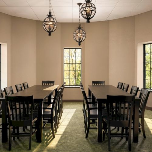 A small, sunlit dining room with two long dark wood tables, about 14 chairs, beige walls, three hanging lantern-style lights, and large windows.